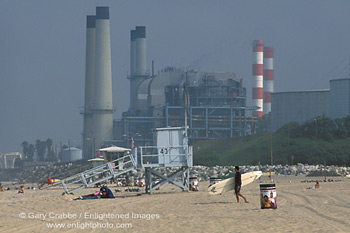 Surfer walking across sand below industrial power plant and smokestack, Manhattan Beach, Los Angeles County, California