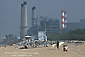 Surfer walking across sand below industrial power plant and smokestack, Manhattan Beach, Los Angeles County, California