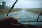 Picture: Hand on steering wheel looking through windshield glass and wipers to a rain soaked country highway, New Mexico